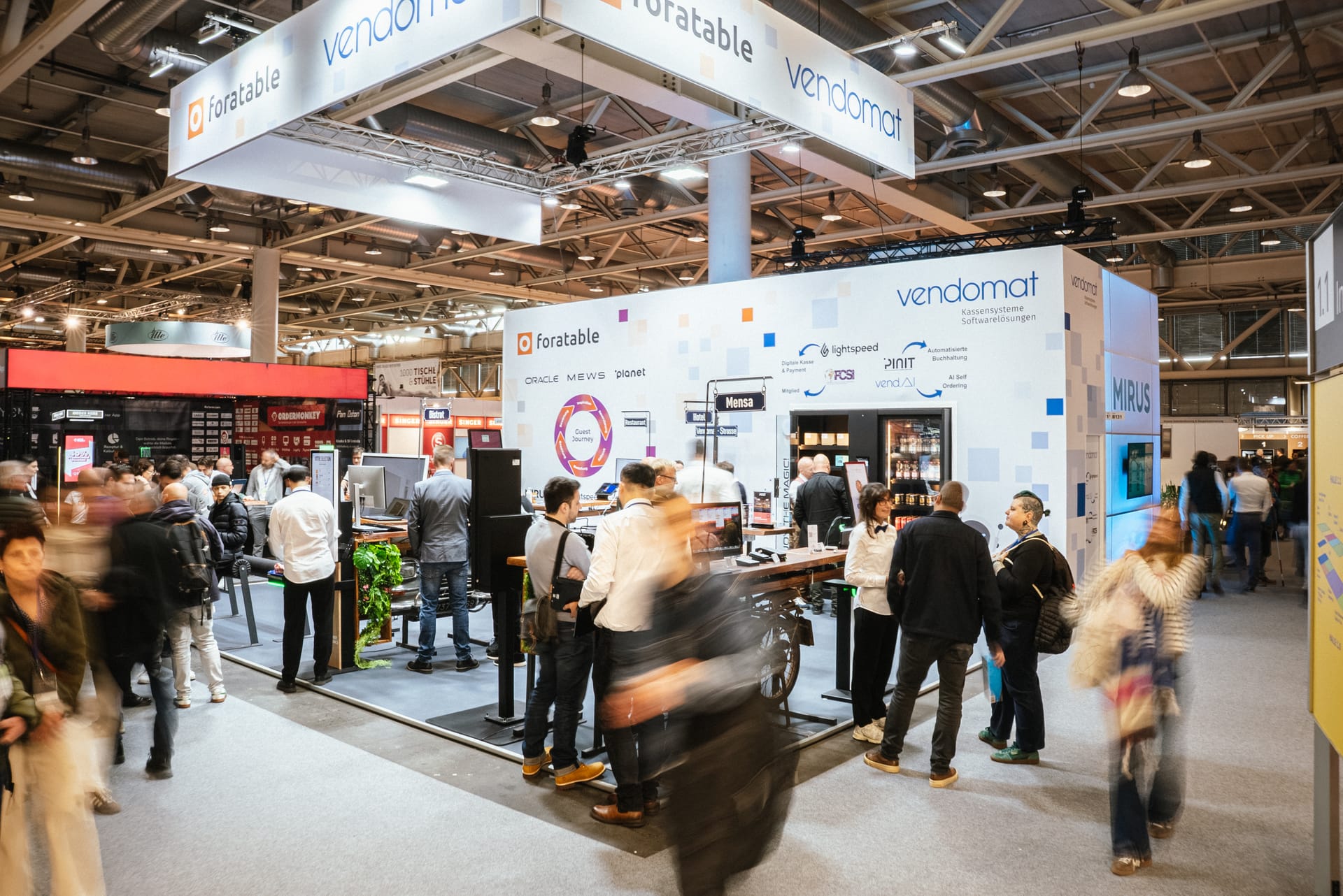 Vendomat Stand an der Igeho Fachmesse mit stark frequentiertem Setup, Partnerzonen wie Foratable und diversen Kassensystem-Demos. Besucher erkunden live die Vendomat Lösungen.