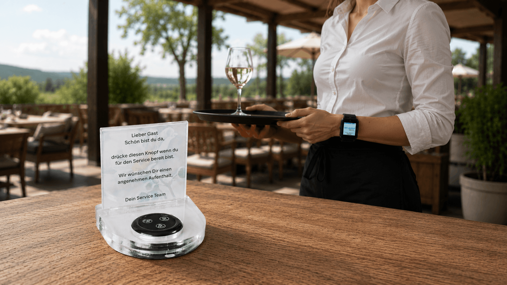 Guestline Service-Aufsteller mit Rufknopf und freundlicher Nachricht für Gäste steht auf einem Holztisch auf einer grosszügigen Restaurantterrasse. Im Hintergrund trägt eine Kellnerin die Guestline Smartwatch mit Vendomat Logo zur direkten Servicebenachrichtigung.